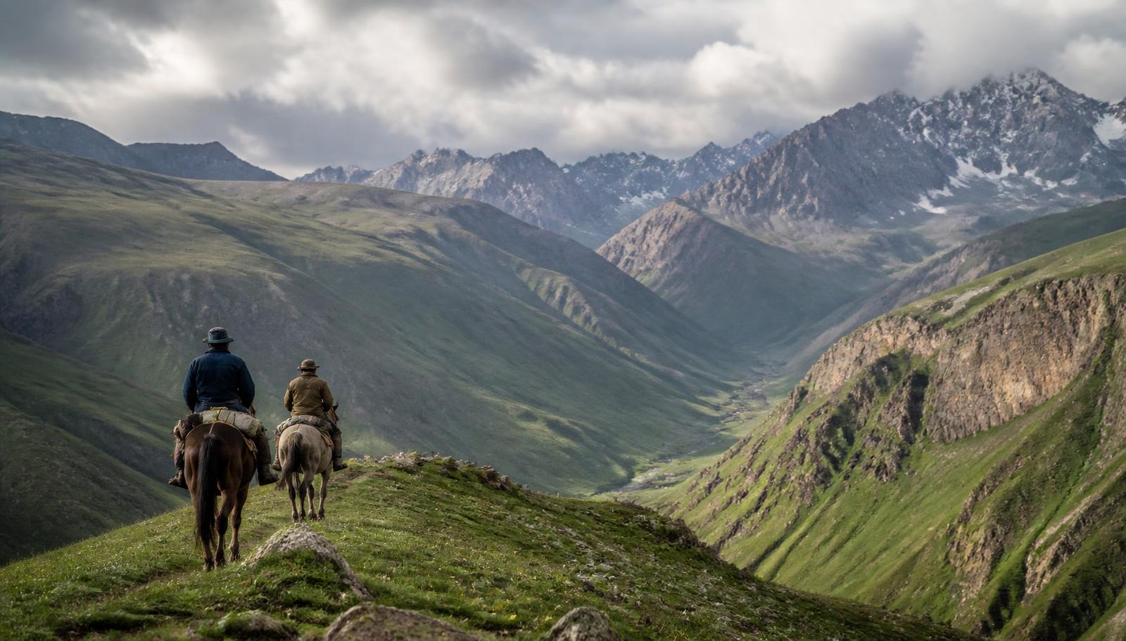 Two riders on horseback crossing a green mountain pass